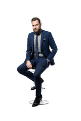 Confident, attractive and
bearded CEO looking elegant in his custom made blue suit and
tie, with one hand in his pocket and sitting on a stool, isolated on
white background.