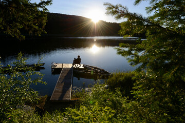Man sitting alone on a wooden chair on a wooden dock/pier/jetty on a lake with kayaks around and the sun shining in the sky and reflected in the water, framed by trees, plants and flowers