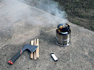 Portable wood burning stove smokes meat on a wooden stick. Nearby lies an ax for splitting firewood...