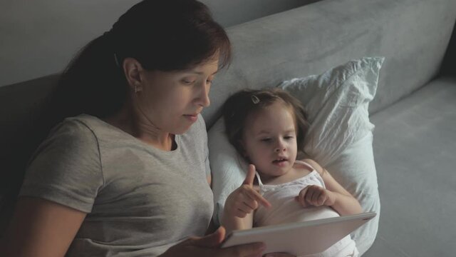 Mom and little daughter are lying in bed and watching a cartoon on tablet. mother and baby make a video call to father. Technological concept, new generation, family, communication, parenthood