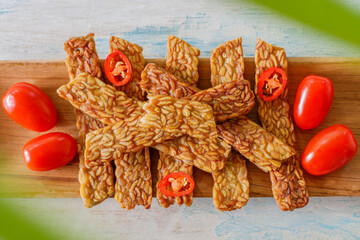 Fried tempeh on a wooden tray over white wooden table, decorated with cherry tomatoes and cut chilly. Top view.