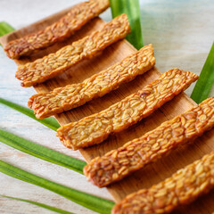 Fried tempeh or tempe on a wooden tray over a wooden table and a green leaf. Top view. Square image.