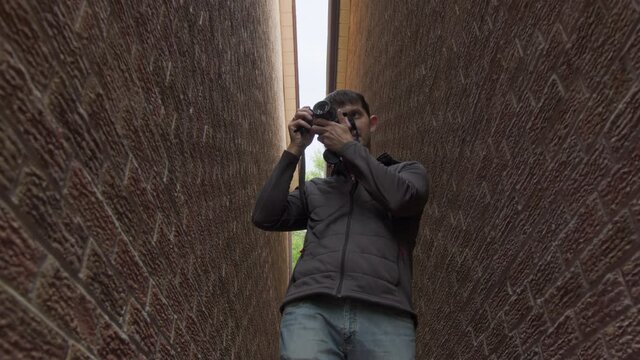 High quality 4k video of a young man who is standing between two red brick walls and holding photo camera in his hands and trying to make a photo or video. Young photographer work in process.