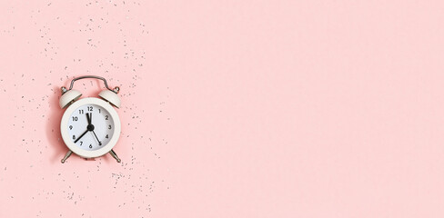 White alarm clock shows about twelve oсlock on pink paper background covered with sequins with copy space. New year Christmas concept. Flat lay. Top view.