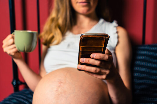 Pregnant woman drinking from mug and using smartphone in bed