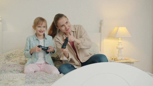 Happy little girl is playing video game with smiling mother relaxing in free time in bed enjoying game. Modern technology, people and house concept.