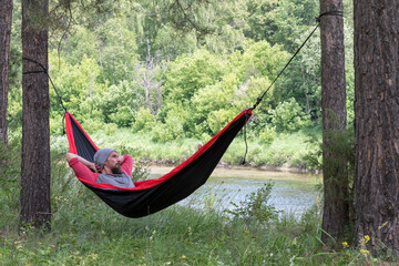 Tourist (man) laying in a hammock on the bank of forest river.