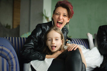 Crazy mother in black leather jaket and pants hugging her little daughter in white dress, they are looking at the camera with opened mouths and grimacing in loft room with houseplants.