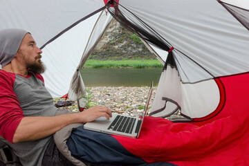 Tourist work on a laptop in a tent on the bank of mountain river while raining.