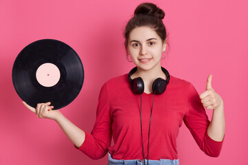 Young adorable girl showing thumb up and holding vinyl music plate in hand, female with knot and...