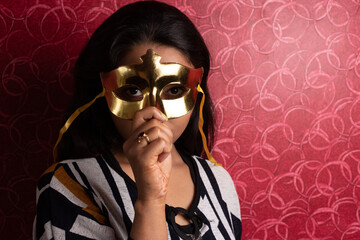 Fashion portrait of an Indian Bengali beautiful and young girl in western dress wearing an eye mask standing in front of a red textured wall. Indian fashion and portrait