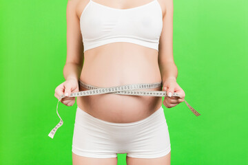 Cropped image of pregnant woman in white underwear measuring her abdomen to check baby development. Centimeter tape measure at green background. Healthy pregnancy. Copy space