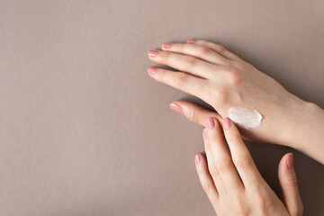 Woman finger applies cream moisturizer on female hand with nude pink manicure on beige background and copy space, above. Beauty skincare concept