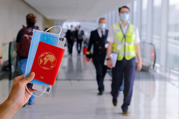 Airport: traveller holding passport, mask, boarding pass, in front of travelator. Airport staff, pilot (wearing face masks) in the background. Travel essential concept; reopening. Selective focus.