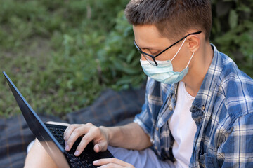 guy in an antiviral mask in a park on a plaid is working on a laptop