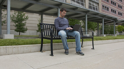 High quality picture of a young men who is sitting on a train station and working on his laptop which is holding on the lap. Young men sitting on a black metal banch on a station in the city. 
