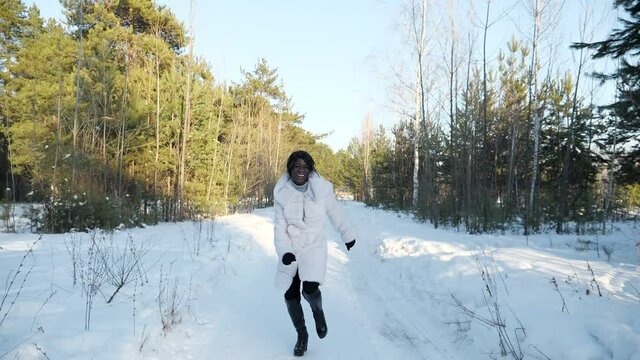 smiling African-American woman in white fur coat runs along snowy path and laughs in winter pine-tree forest slow motion