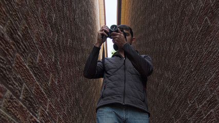 High quality picture of a young man who is standing between two red brick walls and holding photo camera in his hands and trying to make a photo or video. Young photographer work in process.
