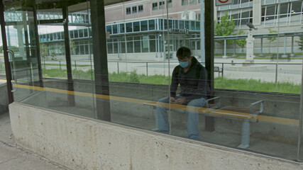 High quality picture of a young man who is waiting for a transportation in a glass bus stop or station and holding his cell phone in his hands. Man is texting on a station while waititng for a train