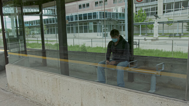 High quality picture of a young man who is waiting for a transportation in a glass bus stop or station and holding his cell phone in his hands. Man is texting on a station while waititng for a train