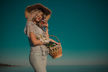 young curly blonde with a glass in hand in a straw hat with daisies in a basket on a sky background