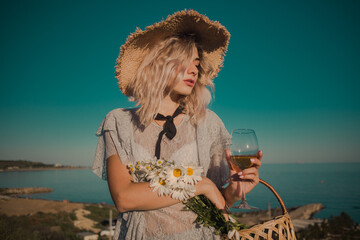 young curly blonde with a glass in hand in a straw hat with daisies in a basket on a sky background