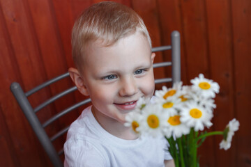 little boy in a white T-shirt sits on a chair against a wooden wall and holds a bouquet of chamomiles in his hands, looks at him and smiles.
lmage with selective focus