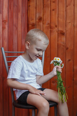 blond boy in a white T-shirt sits on a chair against a wooden wall and holds a bouquet of chamomiles in his hands and smiles.
lmage with selective focus