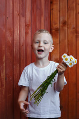 a little blond boy in a white T-shirt stands against a wooden wall and holds a bouquet of chamomiles in his left hand, look into the camera.
lmage with selective focus