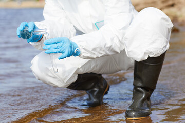 Close-up of chemist in protective workwear and in gloves holding flask and examining the water in...