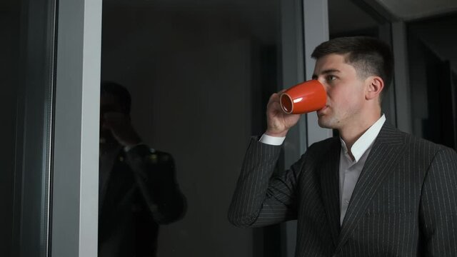 30-year-old businessman in a stylish suit stands in his office near a panoramic window overlooking the night city. He thinks about a business plan and drinks coffee and tea