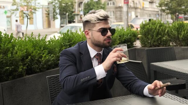 evil businessman smoking cigar and drinking scotch or whiskey in restaurant terrace, elite mens club outdoor. Man straightens hair. man crushes cigar ash