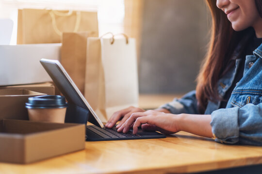 Closeup image of a young asian woman typing on tablet pc for online shopping with postal parcel box and shopping bags on the table at home