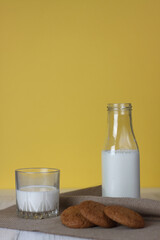 cookies, a bottle and a glass of milk on canvas napkin on white wooden table against yellow background
