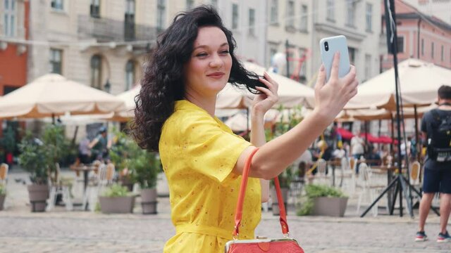 Lifestyle selfie portrait of young positive woman wearing yellow dress having fun and taking selfie in city center smile girl feel happy street smartphone vacation positive fashion close up.