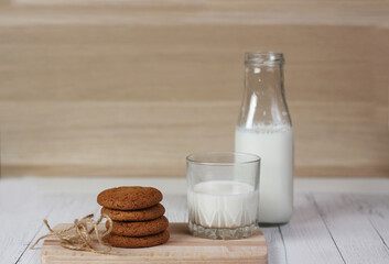 cookies, a bottle and a glass of milk on cutting board on white wooden table against wooden wall
