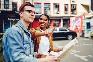 African American young woman inhabitant of city pointing hand on right direction to help Caucasian tourist with map to find destination, friends standing on street and searching showplace in town
