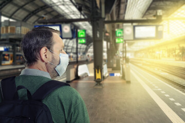 A mature man in face protective mask is photographed on an empty railway platform. A shot from the back on blurred background.