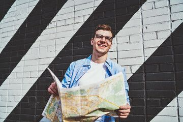 Cheerful caucasian young man in eyeglasses holding travel map in hands and looking away standing in urban setting against wall with graffiti.Happy hipster tourist in casual wear laughing