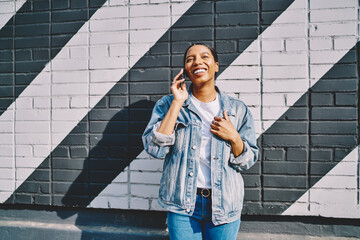 Joyful young woman with dark skin dressed in denim wear and laughing during phone conversation on smartphone.Positive african american hipster girl calling on mobile phone and talking with friend