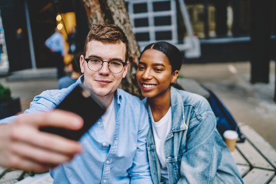 Positive multicultural couple in love spending free time together and making selfie photo on smartphone for social networks.Cheerful diverse friends taking pictures on mobile phone resting outdoors