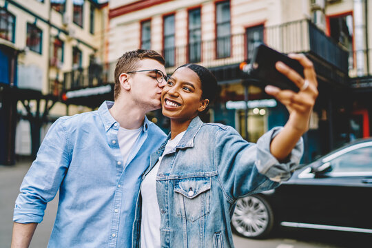 Caucasian hipster guy kissing happy african american girlfriend which holding smartphone and making common selfie during trip in urban setting.Positive diverse marriage taking photo on mobile phone