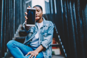 Selective focus on modern smartphone device with black display in african american woman's hands.Blurred background with dark skinned hipster blogger holding mobile phone and showing it at camera