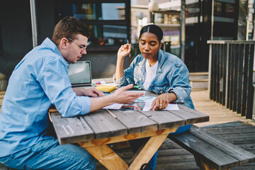 Smart multicultural students sitting at wooden table and discussing solution of studying task during collaboration.Diverse young man and woman cooperating on common project outdoors