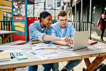 Positive multicultural students dressed in denim wear smiling and watching tutorial on website using wireless internet on laptop computer outdoors.Cheerful diverse designers reading publication