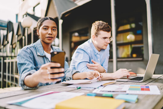 Portrait of african american young woman holding smartphone and chatting online in social networks while caucasian colleague watching tutorial on laptop computer using wireless internet outdoors - Powered by Adobe