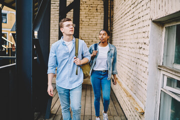 Obraz premium Young multiracial male and female students dressed in jeans trendy outfit walking in college building, teenagers in casual wear strolling with backpacks, hipster couple in stylish youth clothing