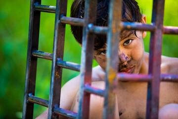 The guilty and angry child sits in a cage with a stern look. Green background outdoors