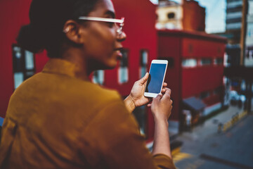 Rear view of African American woman holding smartphone connected to 4G internet standing on street, selective focus on modern mobile phone in hand of dark skinned hipster girl on urban setting