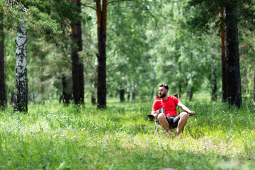 
A young man in a red T-shirt sits in the forest, enjoying the beauties of nature, sits on a folding chair and holds a metal mug